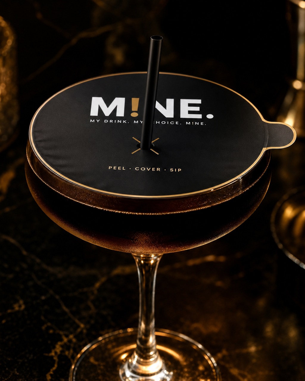 Coupe cocktail glass covered with a matte-black MINE cover with gold outline, pull tab, wordmark, tagline and a straw through the centred gold X opening